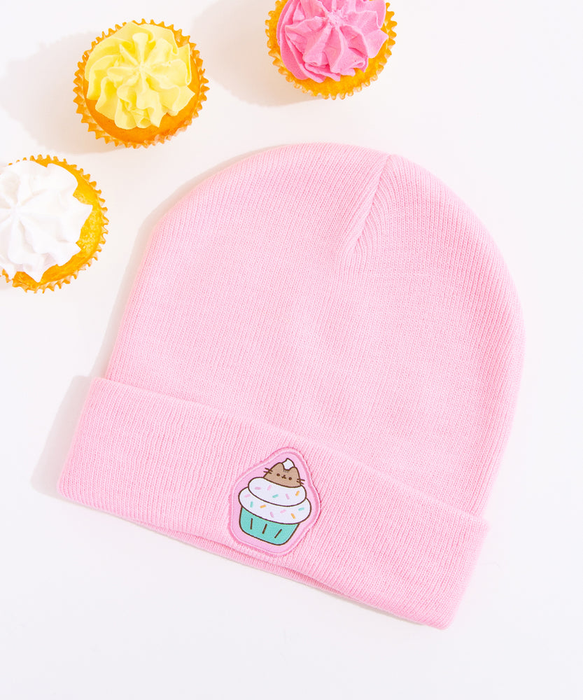 Light pink Pusheen beanie hat laid on a white surface accompanied by cupcakes with white, yellow, and pink frosting. 