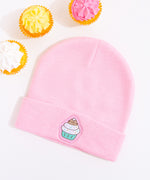 Light pink Pusheen beanie hat laid on a white surface accompanied by cupcakes with white, yellow, and pink frosting. 