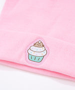 Light pink Pusheen beanie hat laid on a white surface accompanied by cupcakes with white, yellow, and pink frosting. 