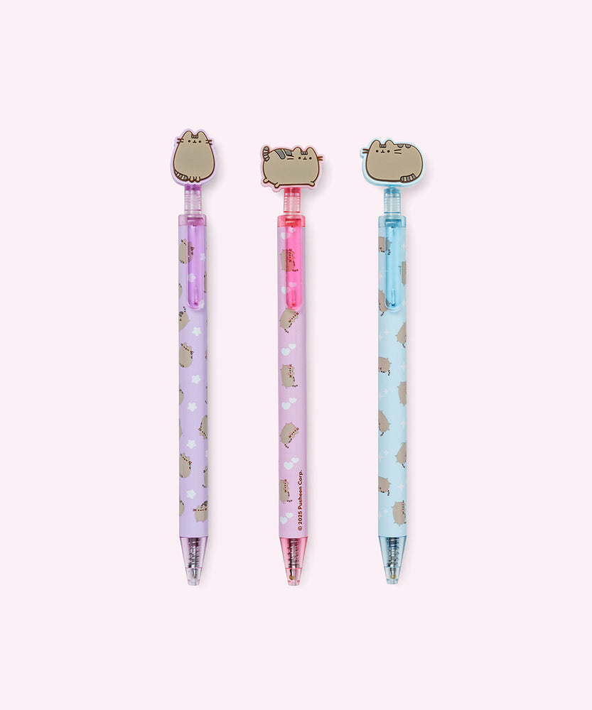 Three pens with Pusheen the Cat tops in a clear holder on a light surface.
