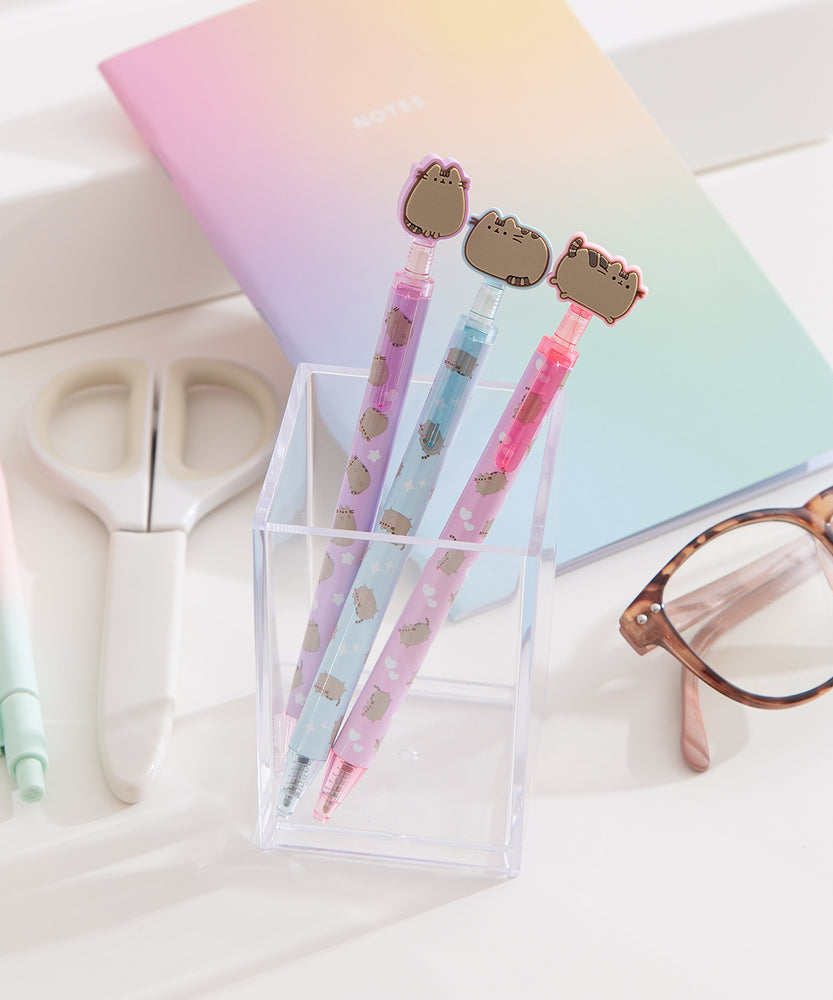 Three pens with Pusheen the Cat tops in a clear holder on a light surface.