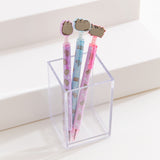 Three pens with cat-shaped ends in a clear holder on a white surface. The pens have purple, blue, and pink outer casings.