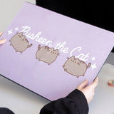 Model holds a purple rectangular desk mat. Across the middle from side to side says "Pusheen the Cat" and features three poses of the gray cat winking, walking, and leaping. 