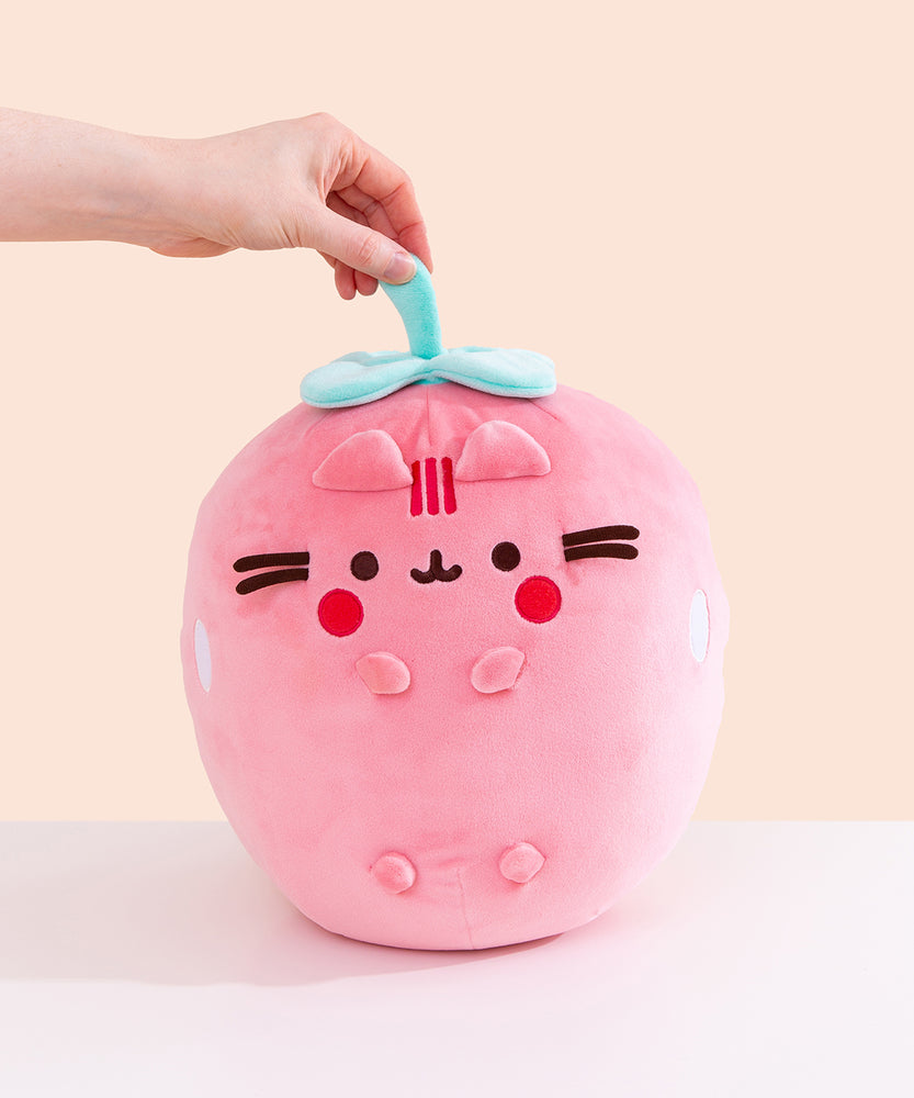 Front view of the Pusheen Fruits Scented Strawberry Squisheen Plush. Pusheen the Cat takes the form of a juicy pink strawberry. The pink body of the rounded plush has white circular shapes on the sides that mimic seeds. On the front of the plush are 3D versions of Pusheen’s ears and four paws. The pink plush is topped with a mint green stem and leaves that a model is holding onto.  