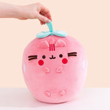 Front view of the Pusheen Fruits Scented Strawberry Squisheen Plush. Pusheen the Cat takes the form of a juicy pink strawberry. The pink body of the rounded plush has white circular shapes on the sides that mimic seeds. On the front of the plush are 3D versions of Pusheen’s ears and four paws. The pink plush is topped with a mint green stem and leaves that a model is holding onto.  