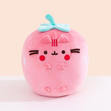Front view of the Pusheen Fruits Scented Strawberry Squisheen Plush. Pusheen the Cat is a light pink strawberry with a mint green stem and leaves that’s sitting atop a white tabletop.  