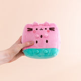 Model holding side of the Pusheen Fruits Watermelon Plush. Pusheen the Cat takes the form of a juicy pink and mint green watermelon slice. The triangle shaped plush body has embroidered brown cylinder shapes to represent watermelon seeds. On the front of the plush are 3D versions of Pusheen’s ears and four paws.  