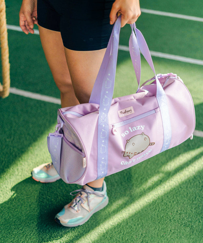 Model holds the top straps on the purple duffle bag. A mesh pocket is located on one side of the bag. Printed on the front of the bag is a graphic of Pusheen the Cat with the phrase "so lazy, can't move."