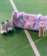 Pusheen Gym Bag sits on a turf surface with a water bottle. The purple bag has a velcro and zippered pocket on one side for easy access storage. 