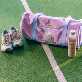 Pusheen Gym Bag sits on a turf surface with a water bottle. The purple bag has a velcro and zippered pocket on one side for easy access storage. 