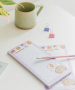 Two purple and white notepads on a desktop. The lined pages include a pink checkbox on each line.