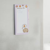 Pusheen Magnetic Shopping List hangs on a stainless steel fridge. The purple and white lined sheets feature graphics of Pusheen and sweet and savory treats. 