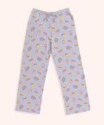 The Snack Time Pajama Pants are laid out to show the full length of the pants. The pants are covered in a pattern of Pusheen, hamburgers, French Fries, and the phrase “YUM” in pink and yellow print. 