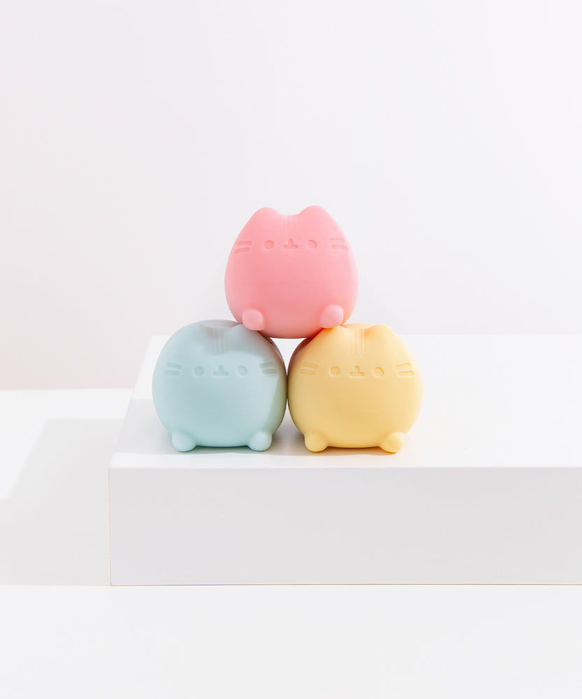 The Pusheen pink squishy toy is stacked onto of the blue and yellow squishy toys that are shaped as a lying down Pusheen the Cat.