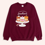 On a light backdrop lies a long sleeve burgundy graphic sweatshirt. The Pusheen Sweets Sweatshirt features a large graphic of Pusheen the Cat as three, layered cakes. The brown mocha cake on the left features chocolate sprinkles and is topped with whipped cream. The middle strawberry cake is layered with yellow cake and features cut strawberries throughout. The right caramel cake features caramel drinks off the top.  