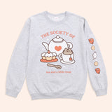 Front and sleeve view of the Pusheen Tea Society Unisex Sweatshirt. The arms are laid out to reveal the printed sleeve detail on the wearer’s left sleeve. The sleeve features rust, cream, and white icons of teacups, biscuits, and a tea kettle. 