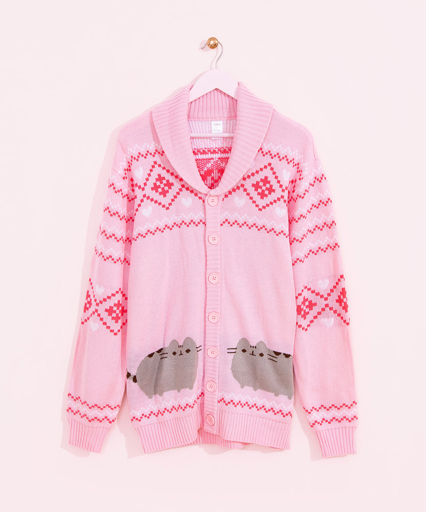 Pink knit cardigan hangs on a pink hanger in front of a light pink wall. The unisex cardigan features Pusheen the Cat in grey and brown surrounded by dark pink diamond pattern and white hearts and diamond pattern all over the sleeves and top of button up cardigan. 