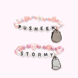 Front view of the Pusheen and Stormy Best Friends Bracelet Set. One of the bracelets has light pink round beads in varying sizes paired with white stars and silver beads with the letters “PUSHEEN” spelled out in beads in the middle and a dangle charm of Pusheen the Cat. The second bracelet has a dangle charm of Little Sister Stormy with purple round beads, pink heart beads, and white small beads. In the middle of the second bracelet is “STORMY” spelled out in letter beads. 