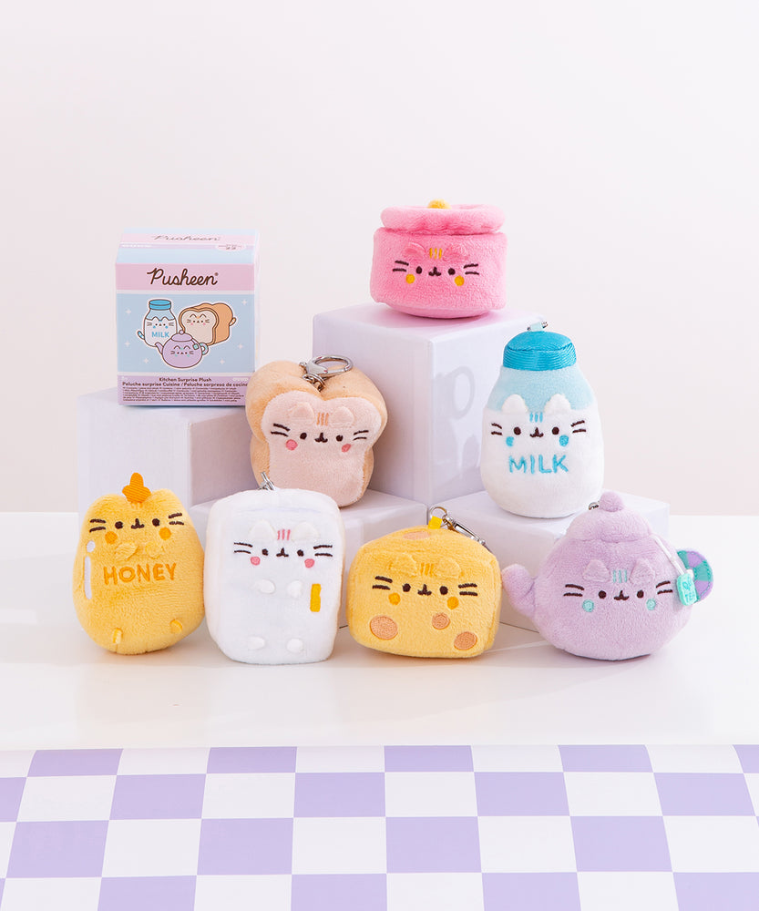 Front view of Pusheen’s Kitchen Surprise Plush assortment. Plush keychains sit on multi-level white pedestal in front of a white background with a purple checkered tabletop. Keychains feature various kitchen essentials including a pot, white bread slice, milk jug, honey pot, mini refrigerator, cheese slice, and tea pot. Accompanying the seven kitchen keychains is the mystery box packaging for the Kitchen Surprise Plush. 