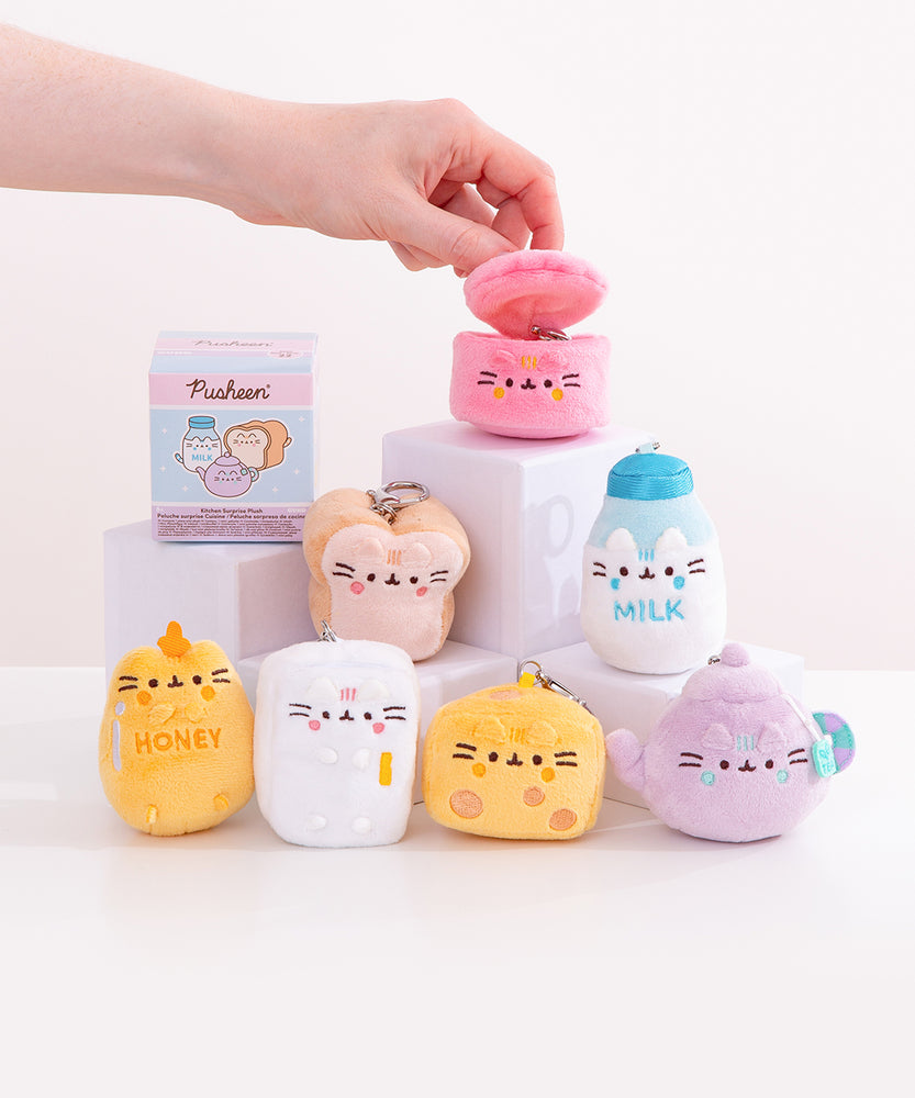 Model holds the top lid open on the pot kitchen keychain. The pink plush is surrounded by a white and blue milk jug, cream and light brown, white bread slice, lilac tea pot, spotted cheese slice, white refrigerator, and yellow honey jar. 