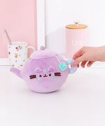 Back view of the Pusheen's Kitchen Teapot Squisheen Plush. Looking at the back, the lilac colored tea kettle lid and pour spout can be seen as well as the mint green and lilac striped handle extending off the round plush body.