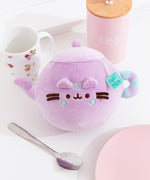 Teapot Squisheen Plush lies on a white tabletop. The lilac plush is surrounded by a tea cup, tea holder, plate, and spoon.