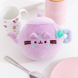 Teapot Squisheen Plush lies on a white tabletop. The lilac plush is surrounded by a tea cup, tea holder, plate, and spoon.