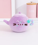 Pusheen's Kitchen Teapot Squisheen Plush sitting on a white surface. Pusheen the Cat takes the form of a lilac tea kettle. One paw extends to become the spout.