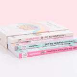 I Am Pusheen the Cat, The Many Lives of Pusheen the Cat, and Pusheen the Cat's Guide to Everything Paperback books stacked atop one another inside the decorative book box. Books lie upside down inside the box to show the spine graphics and portions of the back covers of each book.  
