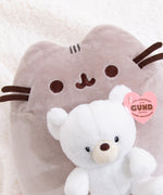 Pusheen x GUND Kai Bear Plush lies on a white fluffy surface. The grey cat holds a white teddy bear in front of her body.