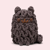 Front view of the Pusheen x The Woobles Pip Crochet Kit. The dark grey cat is crafted with yarn that mimics Pip’s unique shaggy fur pattern. The plush stands on two legs and has a tail feature. 