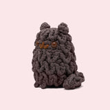Side view of the Pusheen x The Woobles Pip Crochet Kit. The dark grey cat is crafted with yarn that mimics Pip’s unique shaggy fur pattern. The plush stands on two legs and has a tail feature. From this view, the crochet tail can be seen coming out of the bottom back of the figure.