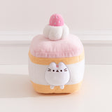 Pusheen Cake Plush is a layered strawberry sponge cake with alternating light yellow cake, white cream, and light pink icing. The cake is topped with a white textured whipped cream plush dollop and light pink strawberry with white embroidery to look like seeds.
