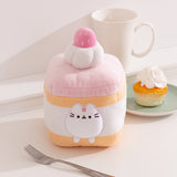 Front view of the Pusheen Sweets Strawberry Spongecake Squisheen Plush. The light yellow and pink Pusheen plush sits within the layers of the cake that's topped with whipped cream and a pink strawberry.