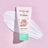 Front view of the Pusheen Strawberry Milkshake Hand Creme. The light pink tube has a printed graphic of Pusheen holding a milkshake surrounded by light pink strawberries. Behind the squeeze tube is a swatch of the white hand cream.