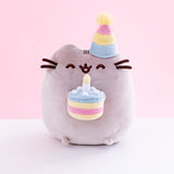 Pusheen sitting upright, eyes closed upwards and mouth open, with a striped party hat over her right ear. Her two nub paws hold a plush birthday cake with a single candle in it, and her bottom nub feet rest at the bottom. The plush is in front of a white and pink background.