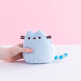 A model’s hand holding the Blue Mini Squisheen Plush in front of a white and pink background. The blue plush has dark blue stripes.