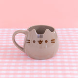 Pusheen Character Mug sits on a pink and white plaid surface. The grey mug has two ears to mimics Pusheen the Cat's face. On the front of the mug, is a printed face with brown whisker, eyes, and mouth details with grey stripes in between the ears.
