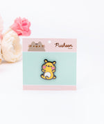 The Cheek pin in it’s packaging, laying on a white floor, a pair of pink and white peonies partially visible in the top left corner. The packaging is a square cardboard backing, a mint square in the middle and pink banners on the top and bottom. The top banner features the cggcw logo, and the bottom banner says ‘Cheek the Hamster Pin’ in white.