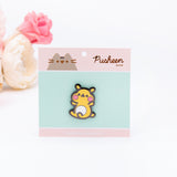 The Cheek pin in it’s packaging, laying on a white floor, a pair of pink and white peonies partially visible in the top left corner. The packaging is a square cardboard backing, a mint square in the middle and pink banners on the top and bottom. The top banner features the cggcw logo, and the bottom banner says ‘Cheek the Hamster Pin’ in white.