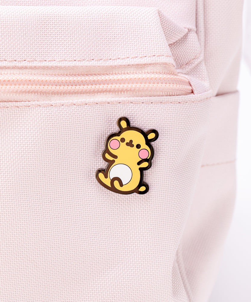 The Cheek pin in it’s packaging, laying on a white floor, a pair of pink and white peonies partially visible in the top left corner. The packaging is a square cardboard backing, a mint square in the middle and pink banners on the top and bottom. The top banner features the cggcw logo, and the bottom banner says ‘Cheek the Hamster Pin’ in white.