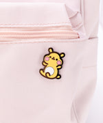 The Cheek pin in it’s packaging, laying on a white floor, a pair of pink and white peonies partially visible in the top left corner. The packaging is a square cardboard backing, a mint square in the middle and pink banners on the top and bottom. The top banner features the cggcw logo, and the bottom banner says ‘Cheek the Hamster Pin’ in white.