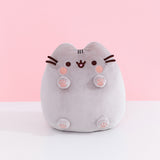 Front view of Classic Toe Beans Pusheen Plush. A blushing Pusheen the Cat shows off her pink toe beans and feet pads on her four paws that extend off her body. Her grey body is accentuated with her classic brown eyes, mouth, and whiskers.  