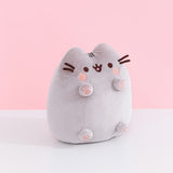 Right quarter view of Pusheen Toe Beans Plush. In this view, Pusheen’s whiskers and feet are shown extended from the plush body.  The plush sits atop a white table in front of a light pink backdrop.