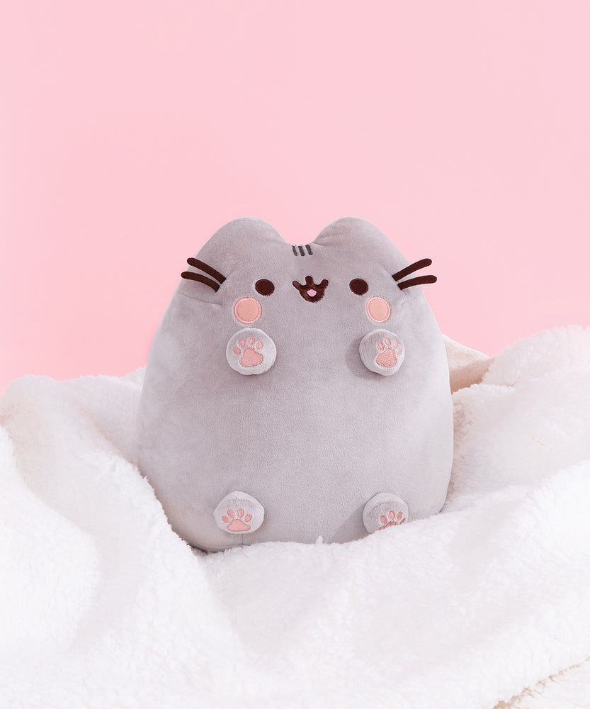Classic Toe Beans Plush sits atop a white fluffy blanket in front of a light pink backdrop. Pusheen’s paws show four light pink paw pads and a large center paw pad. 