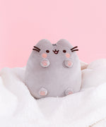 Classic Toe Beans Plush sits atop a white fluffy blanket in front of a light pink backdrop. Pusheen’s paws show four light pink paw pads and a large center paw pad. 