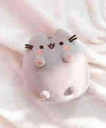 Toe beans plush lays on a white fluffy surface while the sun shines on half of the plush, highlighting the light grey coloring of the plush. Pusheen’s brown whiskers extended off the front side of the plush. 