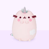 The Dreamy Pusheenicorn Squisheen in front of a light purple background. The plush features a light pink Pusheenicorn sitting upright with a unicorn horn in between her ears, circular blush marks, and an embroidered cloud on the lower right of her front body. Dreamy Pusheenicorn has two nub paws up in the air and two nub paws at the bottom. The embroidery on her eyes, mouth and felt whiskers are a dark magenta, while her head stripes are embroidered in pink.