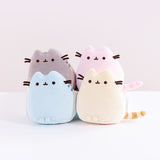 A quartet of Mini Squisheen Plush, all in the same pose with Pusheen in a more triangular shape, resting her feet at the bottom and her tail sticking out to the side. Each plush is a different color, including Pusheen’s classic grey, pink, blue and yellow.