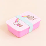 The Hello Kitty x Pusheen Lunchbox closed, with the teal band keeping the lunch box closed. The lid is a lighter pink then the box, and the lunch box is a rounded rectangle. The lunchbox is placed in front of a cream background.